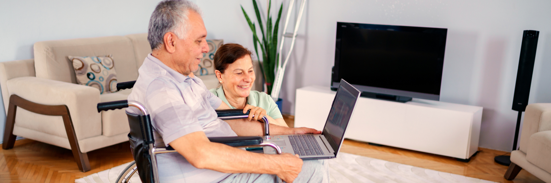An older white man seated in a wheelchair at home talking on a video call with a laptop. A smiling older white woman is kneeling beside him.