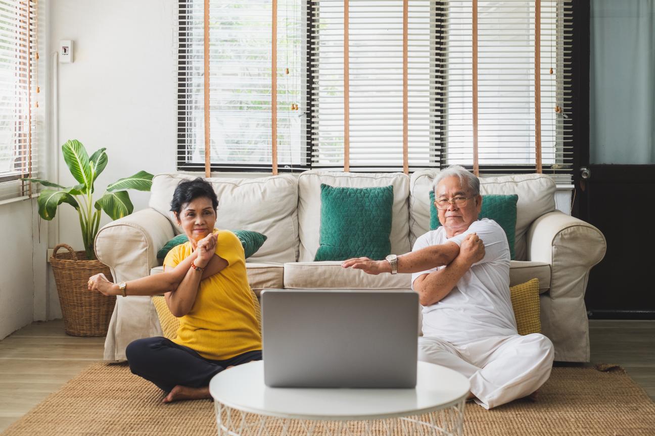 Two older Asian adults doing a seated workout exercise at home with a laptop