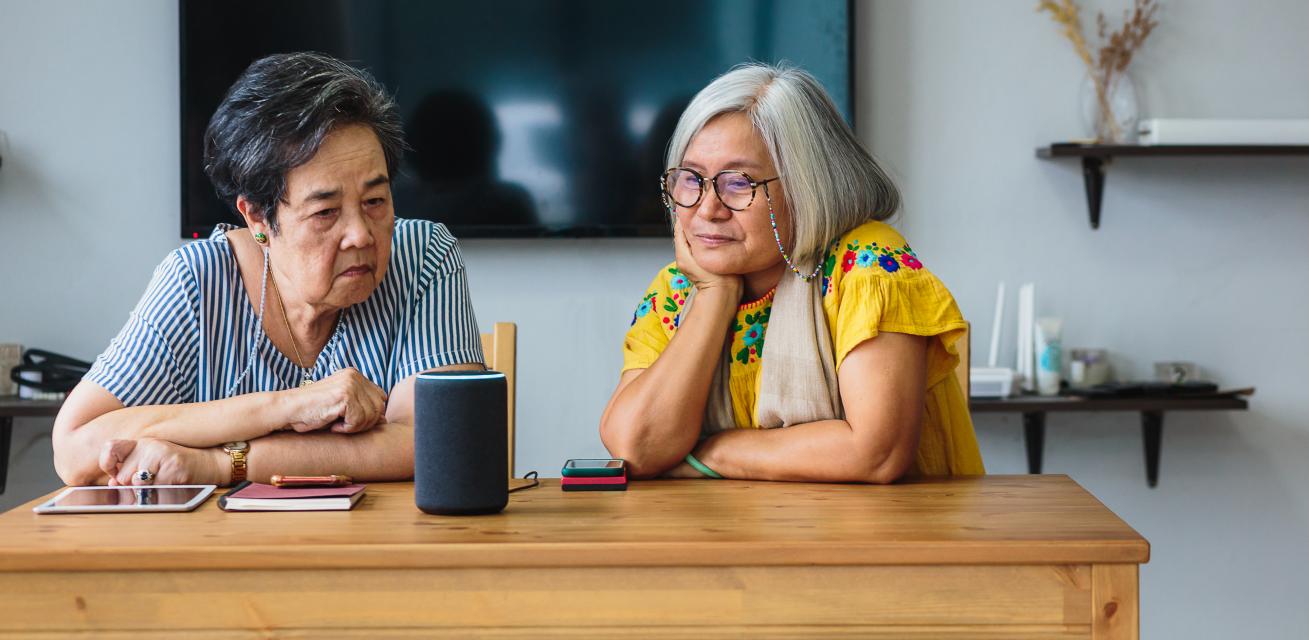Two older Asian women using a smart speaker and electronic tablet while seated at a table at home.