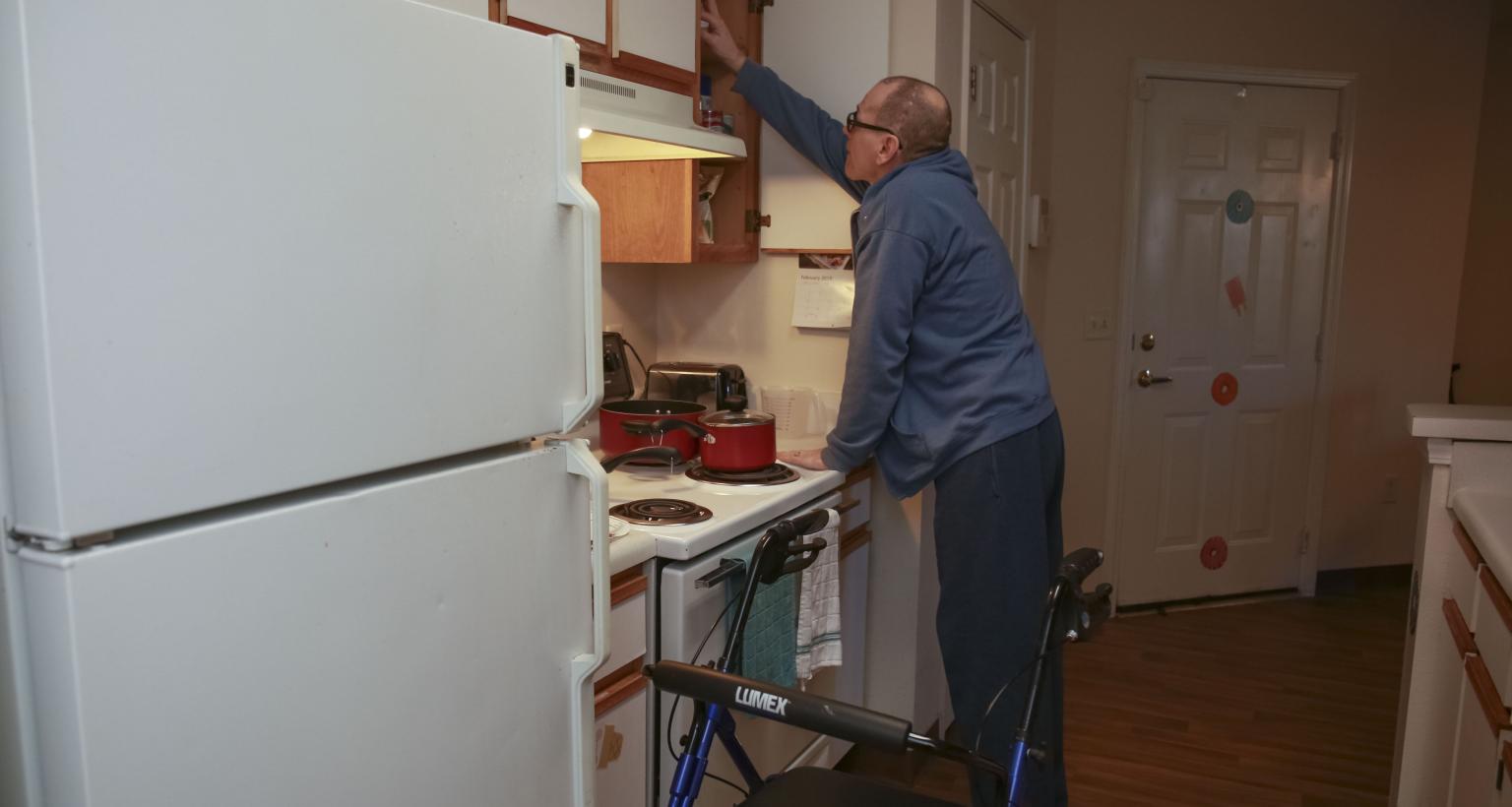 older male who uses walker reaches up into kitchen cupboard