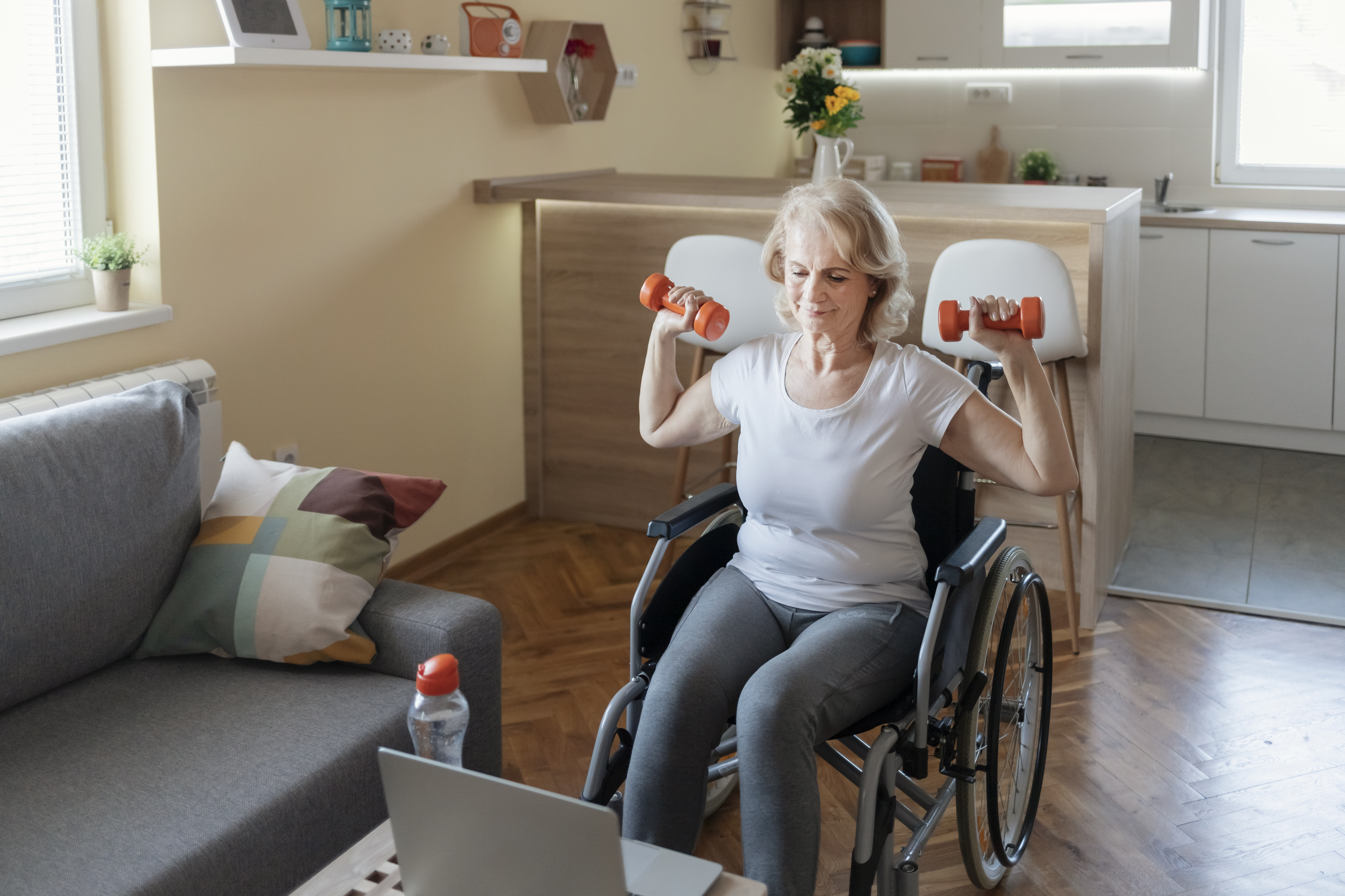 older woman in a wheelchair doing fitness exercises with weights on her computer